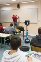 Photo montrant un professeur en train d'écrire au tableau devant une classe de lycéens, avec la présence du robot de téléprésence au premier rang à côté du bureau d'un élève