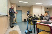 Photo montrant un professeur en train d'interroger des lycéens pendant son cours, avec la présence du robot de téléprésence au premier rang à côté du bureau d'un élève